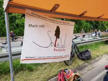 Das Solawi-Banner im Vordergrund, die Mobilitätsspur mit Fahrradfahrer*innen im Hintergrund.
